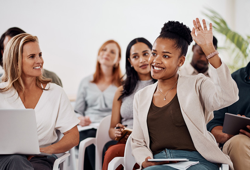 A smiling black woman in her late 20s or early 30s is seated in crowd with her left hand raised. Her hair is pulled back in a bun and she is wearing business casual clothing.