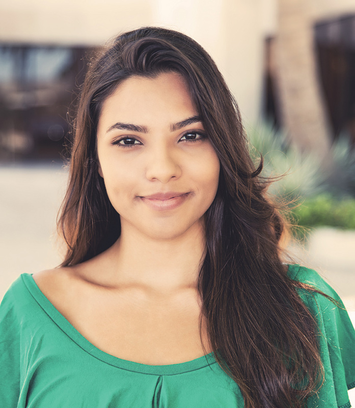 Young Latina woman with light brown skin and long dark brown hair. She is wearing a green blouse and photographed from the waist up. She has a slight grin on her face.