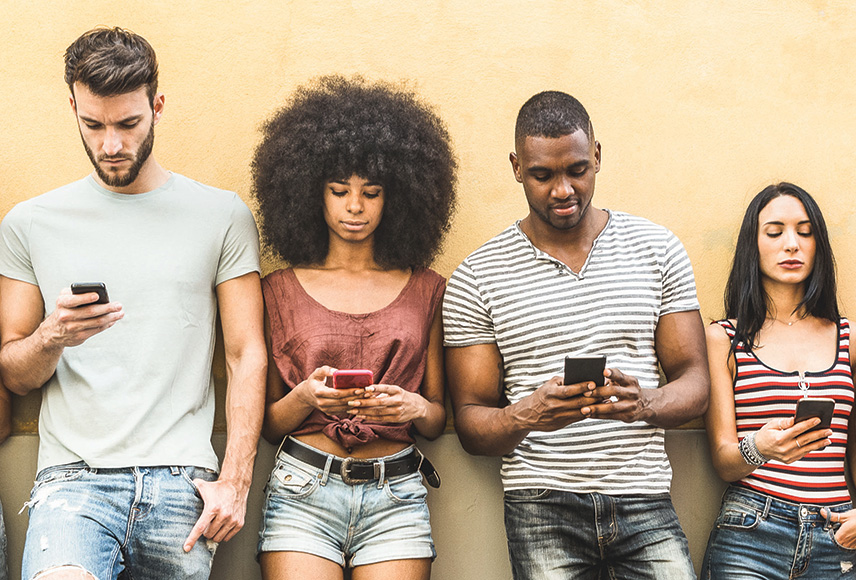 A diverse group of 20-somethings leaning against a yellow wall. Each person is holding a smart phone and looking down at respective screens.