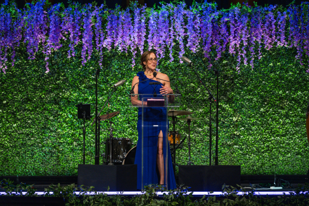 A woman standing at a podium wearing a blue dress, giving a speech with an ivy-draped wall behind her.