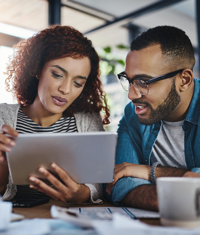 A Black woman and man in their 40s are seated at a table looking at tablet together.