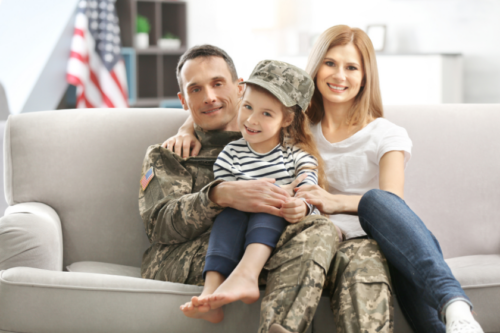 A man in a military uniform sits next to his wife, with their daughter on his lap wearing his hat