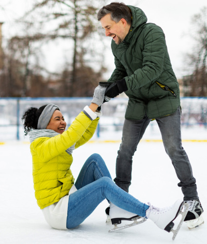 A playful couple on an outdoor ice skating rink. A mixed race woman is seated on the ice. She is wearing a lime green winter coat and white figure skates. The man is white and standing on hock skates in a dark green winter coat. The man is reaching down to help the woman up. Both are grinning and not looking at the camera.