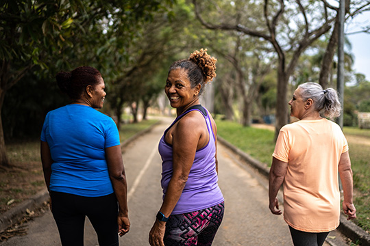 Three women in their 40s and 50s are walking outdoors away from the camera on a paved path. The woman in the middle is wearing purple exercise clothes and is turning her head back. She is making eye contact and grinning.