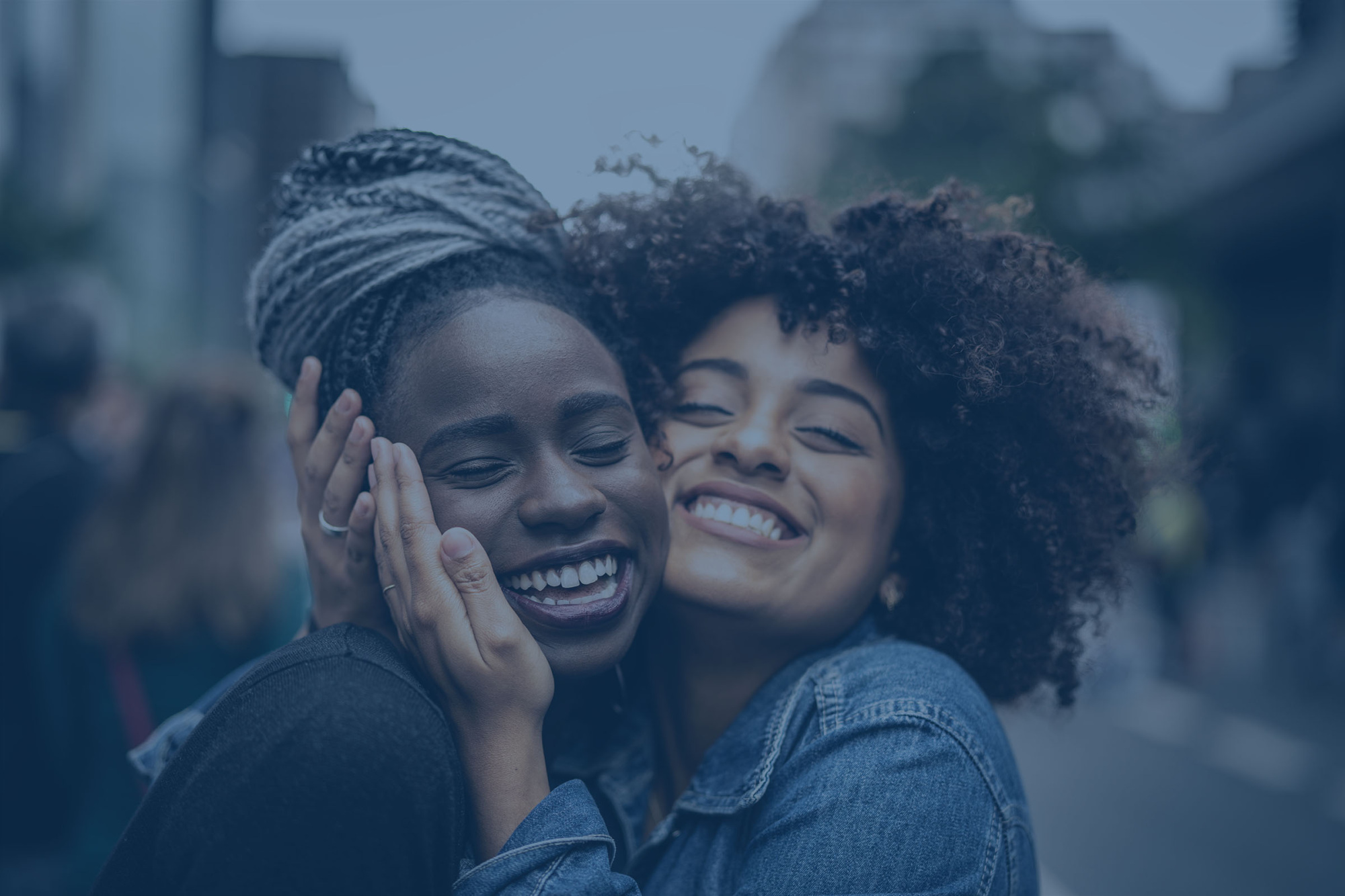 A close up portrait of two Black woman with their heads close together, side by side. Their eyes are closed and they are smiling. The woman on the right is cradling the other woman's face.