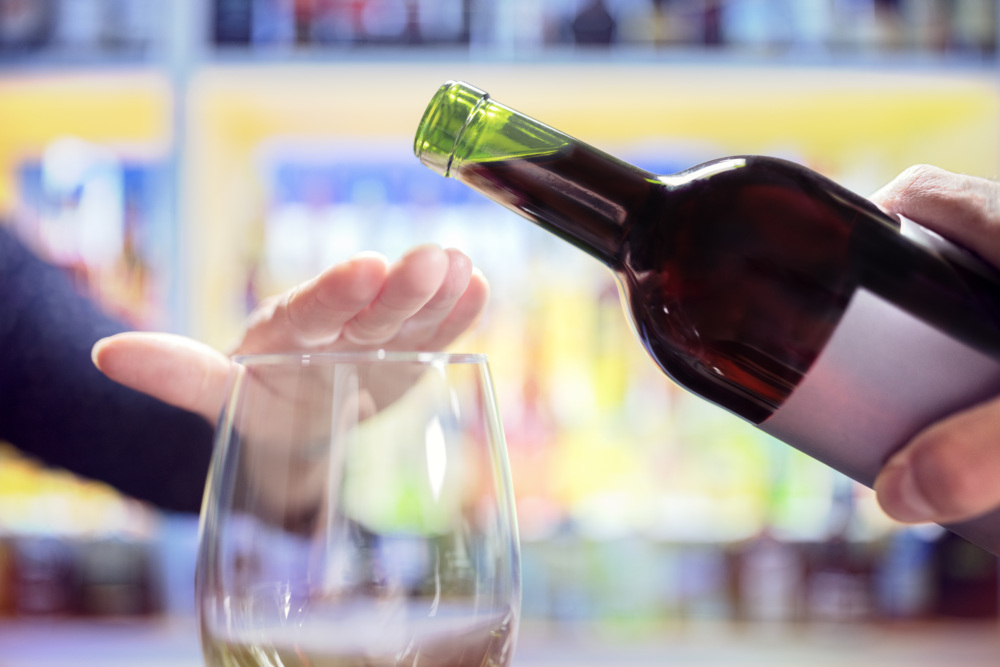 A person puts their hand over their glass to reject the wine bottle that is tipping toward it