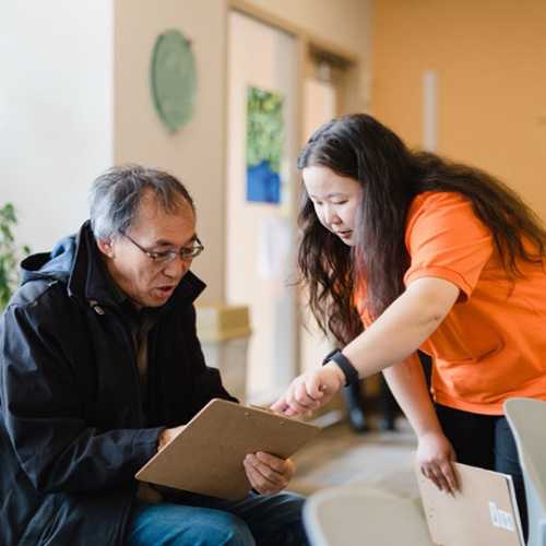 A senior man of Asian decent is seated and reviewing paperwork on a clipboard. A woman of Asian decent in her 20s stand next to the man and is pointing to something on the clipboard.