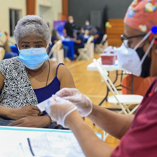 A senior Latina woman is seated in a clinic. She is wearing a mask and is about to get a vaccine. A health care provider is seated next her just out of focus and preparing a bandage.