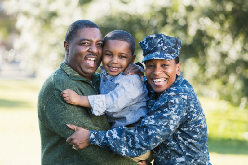 A woman in a military uniform hugs a man holding a young boy