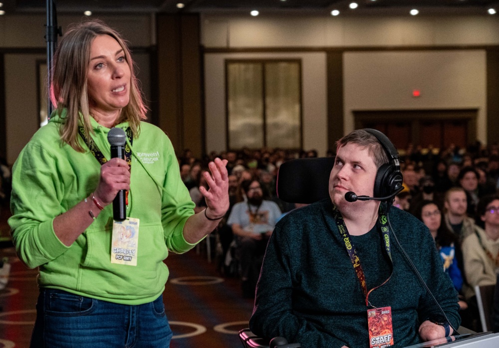 Woman holding up a microphone speaking on a stage with a man in a headset looking at her.