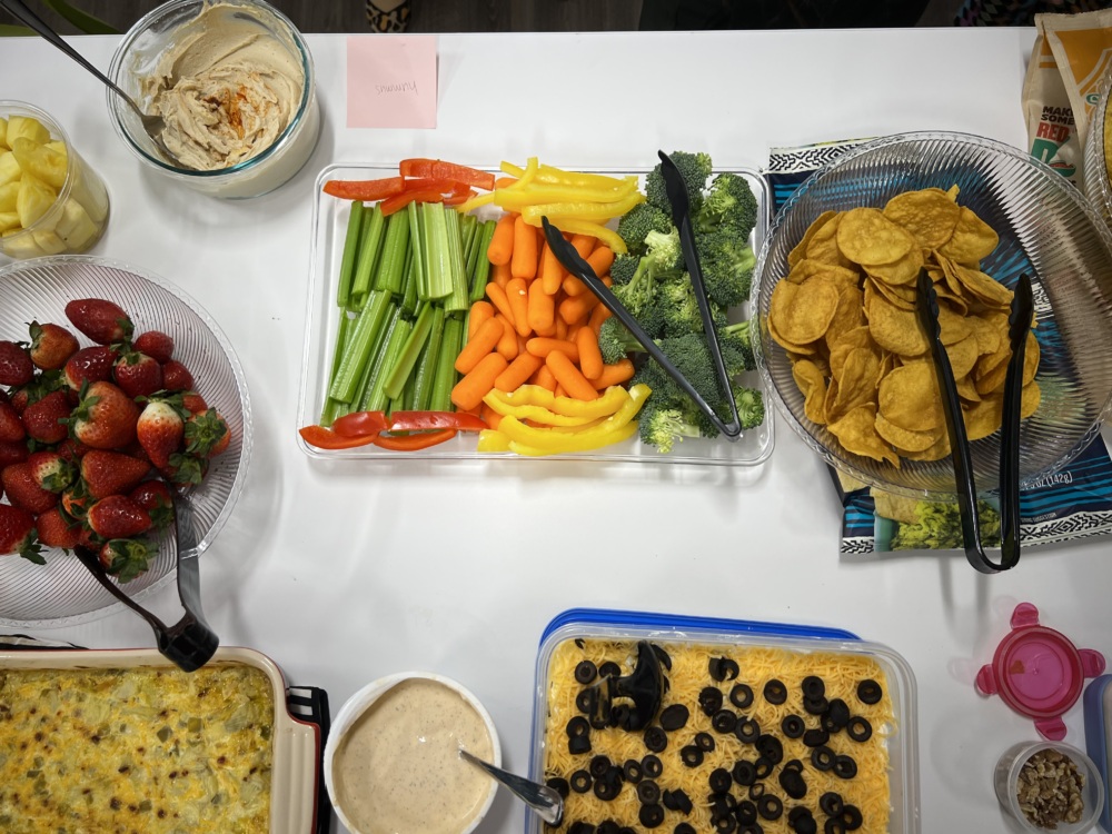 A table with hummus, fruit, vegetables, tortilla chips, artichoke dip and bean dip