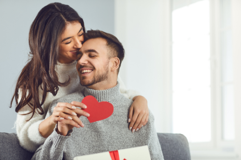 A man holds a paper heart and a gift and a woman has her arms around him