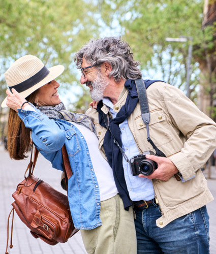 A white woman and man in their 50s or 60s face each other and smile. The main is holding the woman's back and slightly dipping her like in a dance. They appear to be on vacation in the spring.