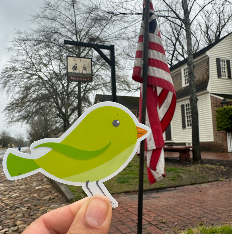 A hand holding a bright green bird cutout in front of an American Flag in Williamsburg, Virginia.