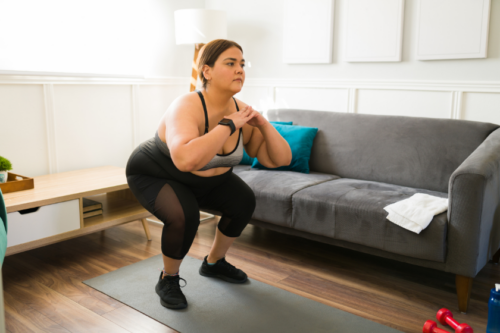 A woman in exercise clothes does a squat on a workout mat in her living room