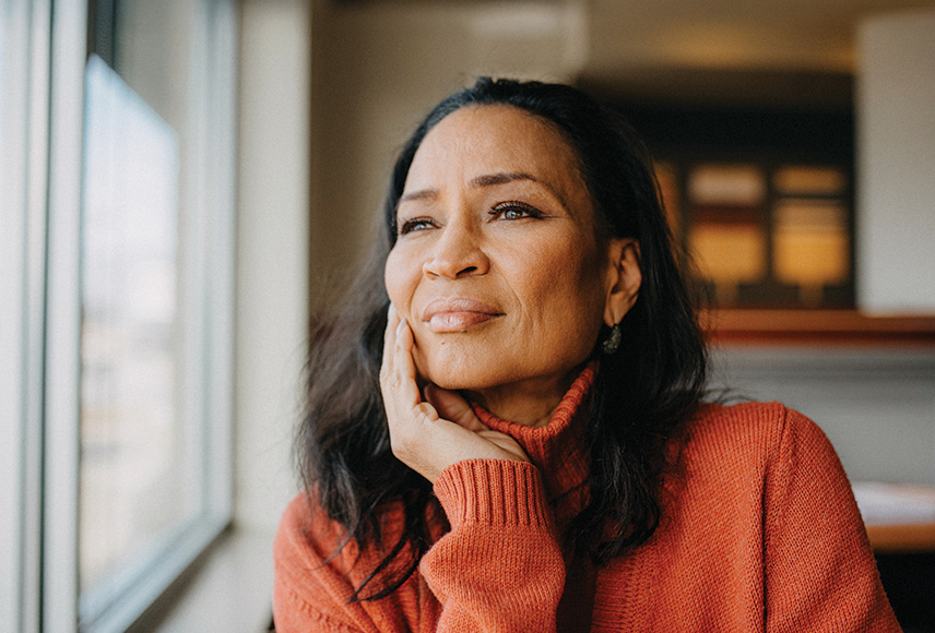 Older Black woman resting her head on her right hand looking thoughtfully out a window.