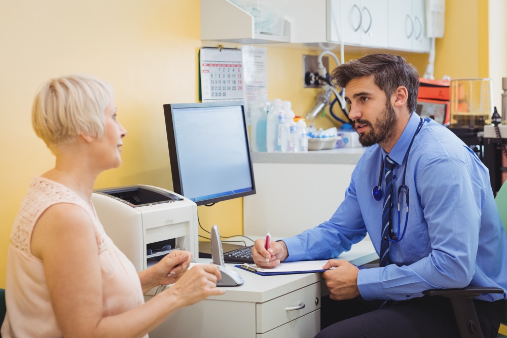 A doctor sits at a desk with a clipboard, speaking to an older woman in a medical office with medical supplies behind them.