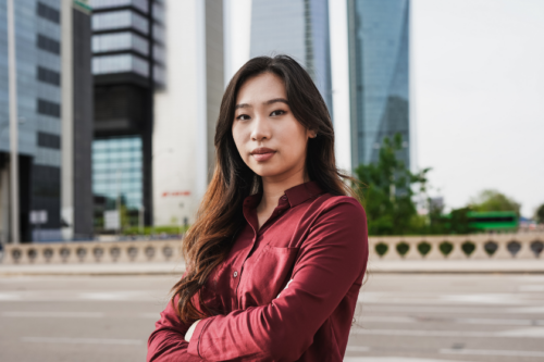 An Asian woman with a serious expression crosses her arms with skyscrapers blurred in the background