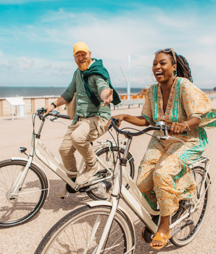 A white man with red hair and beard and a Black woman with long braids are riding bikes together on a sunny beach road.