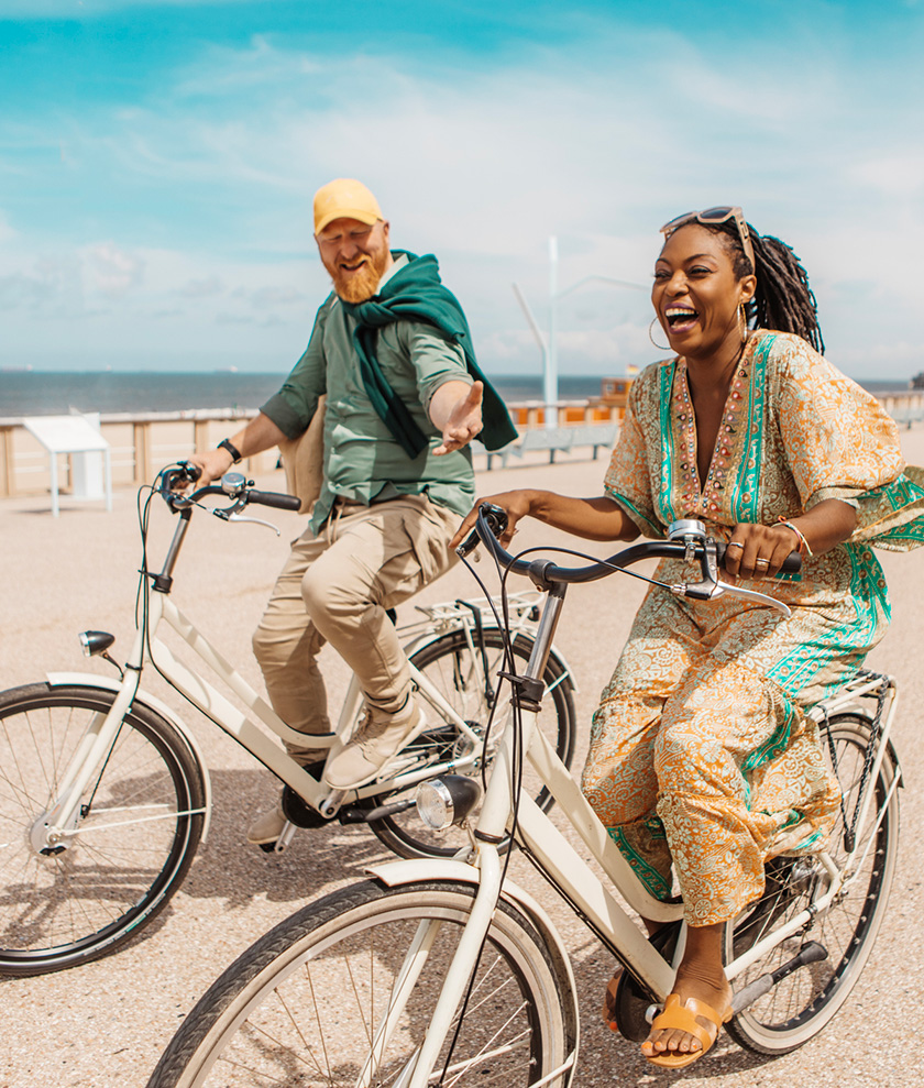 A white man with red hair and beard and a Black woman with long braids are riding bikes together on a sunny beach road.