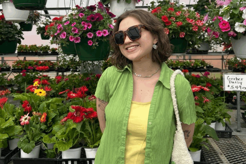 A woman stands smiling for a picture inside a greenhouse with red and yellow flowers behind her.