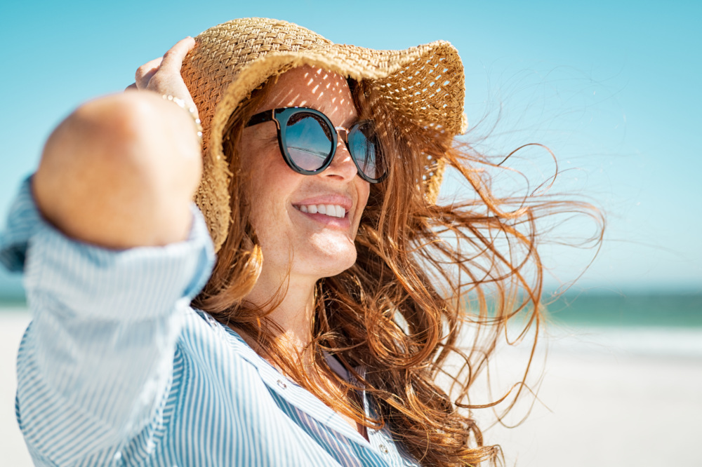 A woman with a floppy straw hat and sunglasses has her brown hair blow in the wind while standing on the beach.