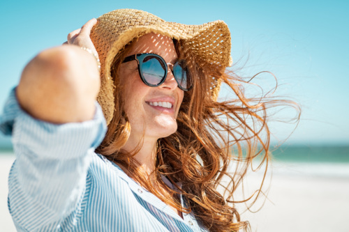 A woman with a floppy straw hat and sunglasses has her brown hair blow in the wind while standing on the beach.