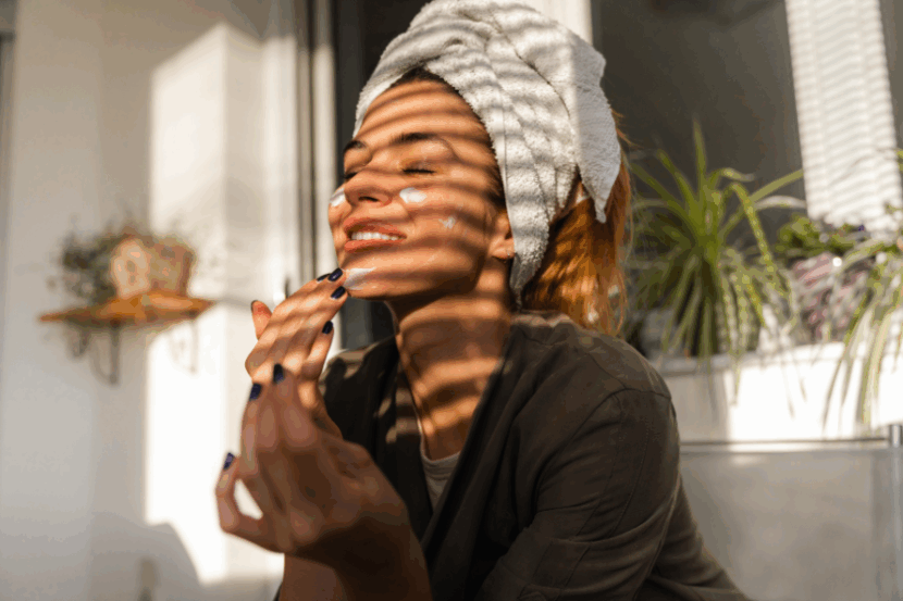 A young woman with a towel wrapped around her hair applies face cream while the sun shines on her.