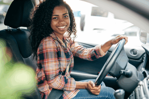 A woman wearing a plaid shirt and a seatbelt smiles in the driver's seat of a car.