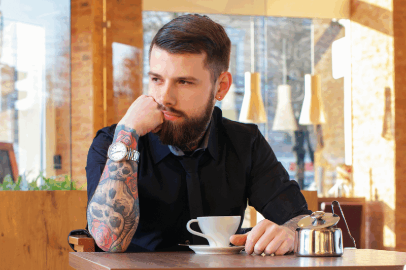 A man with a tattoo sleeve of skulls and roses sits in a cafe with a cup of espresso.