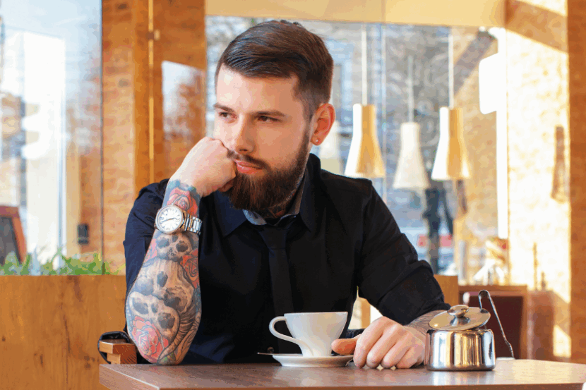 A man with a tattoo sleeve of skulls and roses sits in a cafe with a cup of espresso.