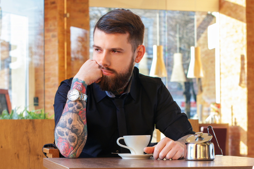 A man with a tattoo sleeve of skulls and roses sits in a cafe with a cup of espresso.