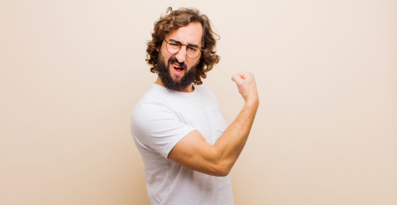 Young bearded crazy man feeling happy, satisfied and powerful, flexing fit and muscular biceps, looking strong after the gym against flat color wall.