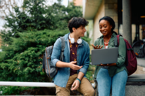 Two college aged students sitting on a wall/pole, looking at a computer screen and smiling.
