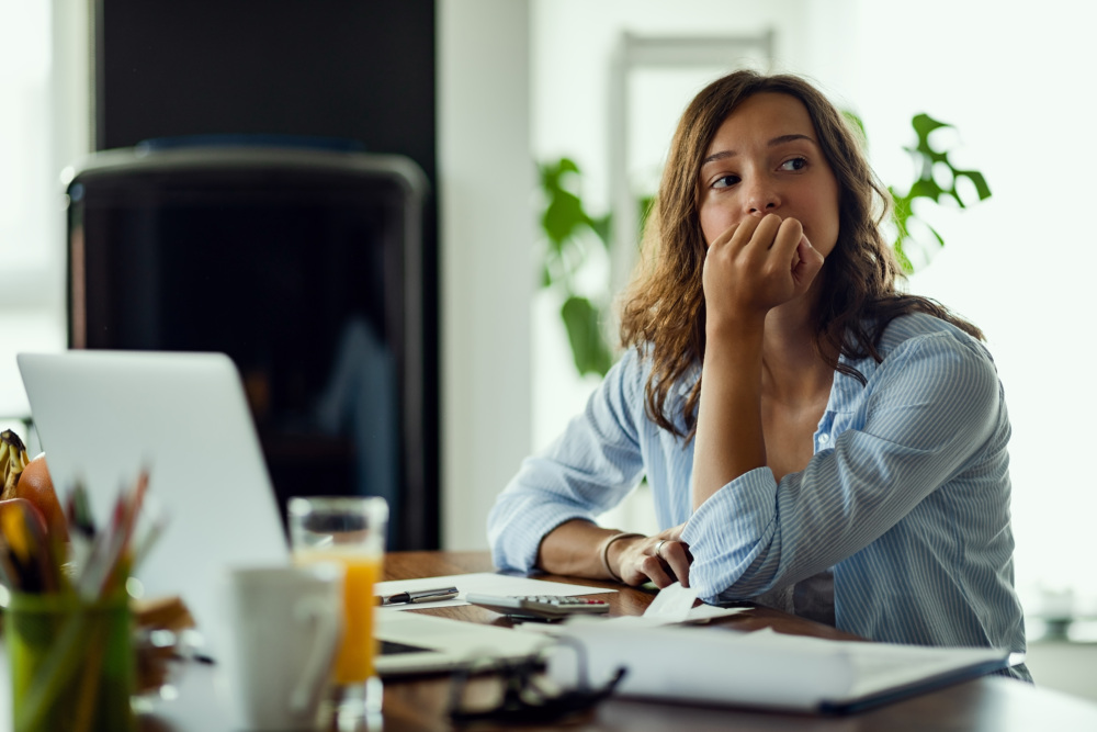 A woman sits at a desk with her hand on her mouth, with a look of concern on her face.