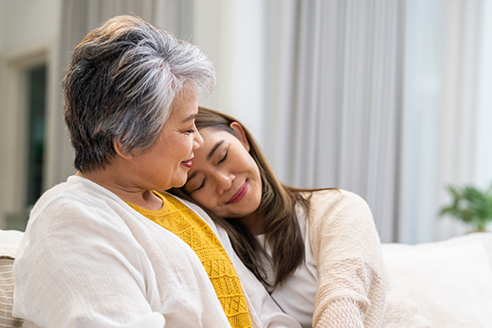 A grandma sits on a couch with her granddaughter who is resting her head on the grandma's shoulder and smiling.