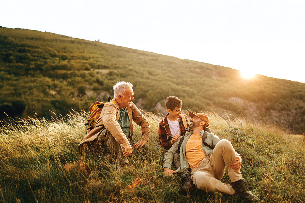 A senior man with his adult son and teen grandson are sitting on a grassy hill while a sun sets behind them.