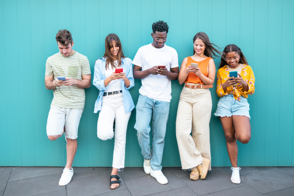 Five people stand against a wall, each looking at their own cell phone.