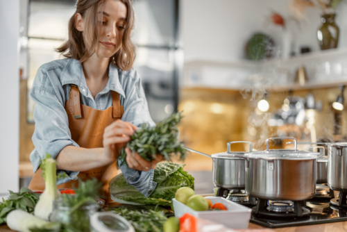 Woman bundles greens together over a kitchen counter next to a pot on the stove.