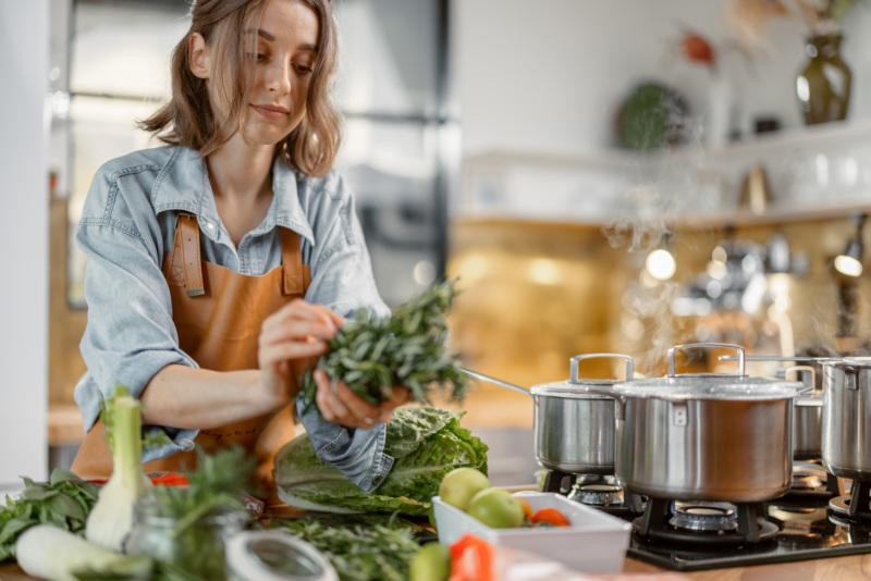Woman bundles greens together over a kitchen counter next to a pot on the stove.