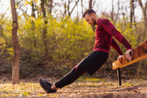 Active man doing bodyweight dips while working out outdoors in street workout park on a sunny autumn day.