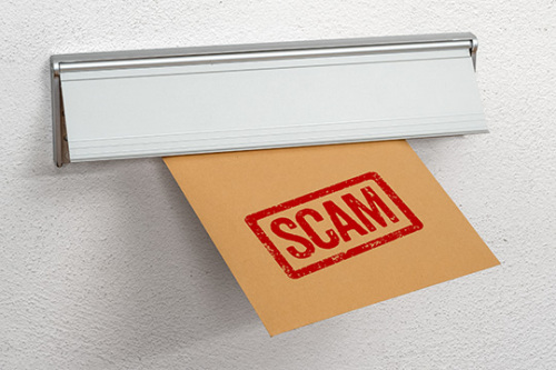 Close up image of a mail slot on the back of a door. A manila envelope is coming through the slot with SCAM in red.