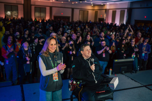 A woman and a man in a wheelchair present on stage while hundreds of people stand behind them, clapping.