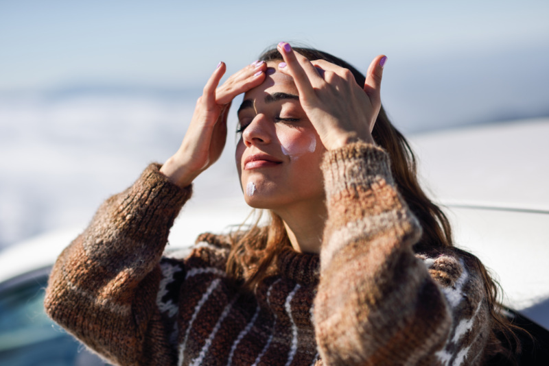 Woman in a sweater outside applying sunscreen to her face.