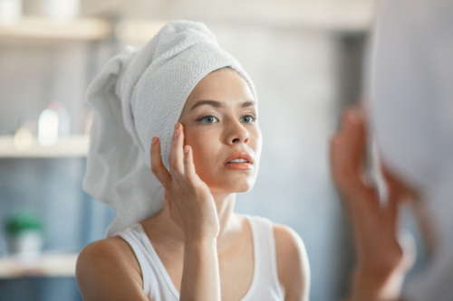 Woman checking her skin in the mirror with a towel on her head.