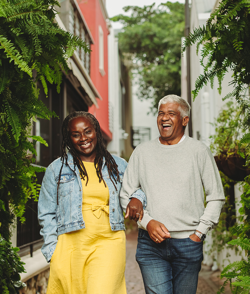 A Black woman and man in their 50s or 60s are walking down a street surrounded by greenery. They are laughing and their arms are linked.
