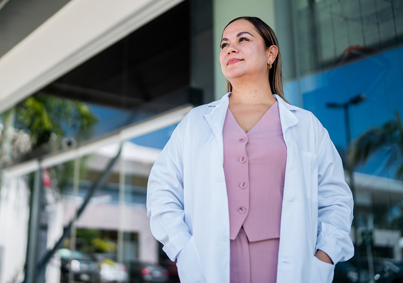 A women in her 30s is standing in front of an institution looking out and with a hopeful expression. She is wearing a white lab coat.
