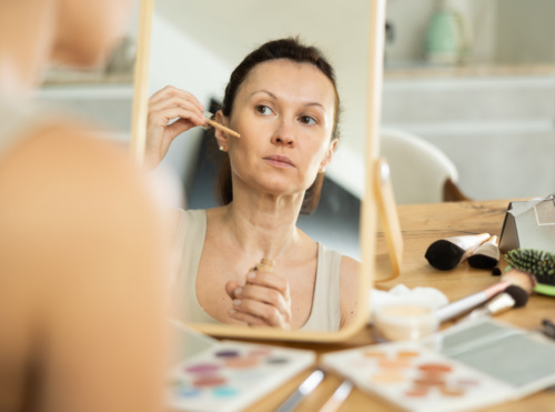 Adult woman starting day with beauty routine in serene home setting in front of vanity mirror, applying cosmetic foundation on facial skin.