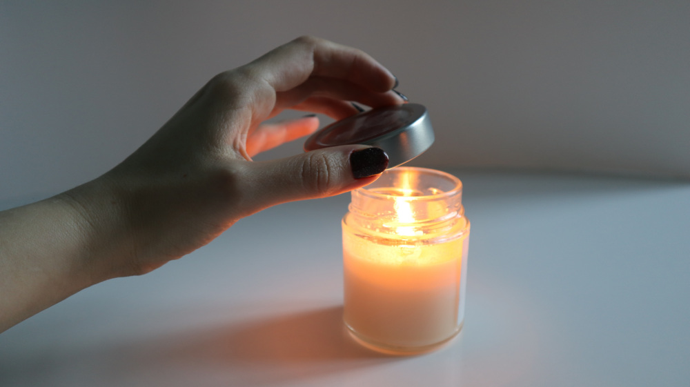 Womans hand covers a burning scented candle with lid, extinguishing the flame. The candle's light creates warm, cozy atmosphere. Idea for meditation, spa procedures, self care, setting romantic mood.