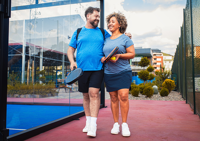 A couple in their 40s walk away from a tennis court smiling and holding pickle ball rackets.